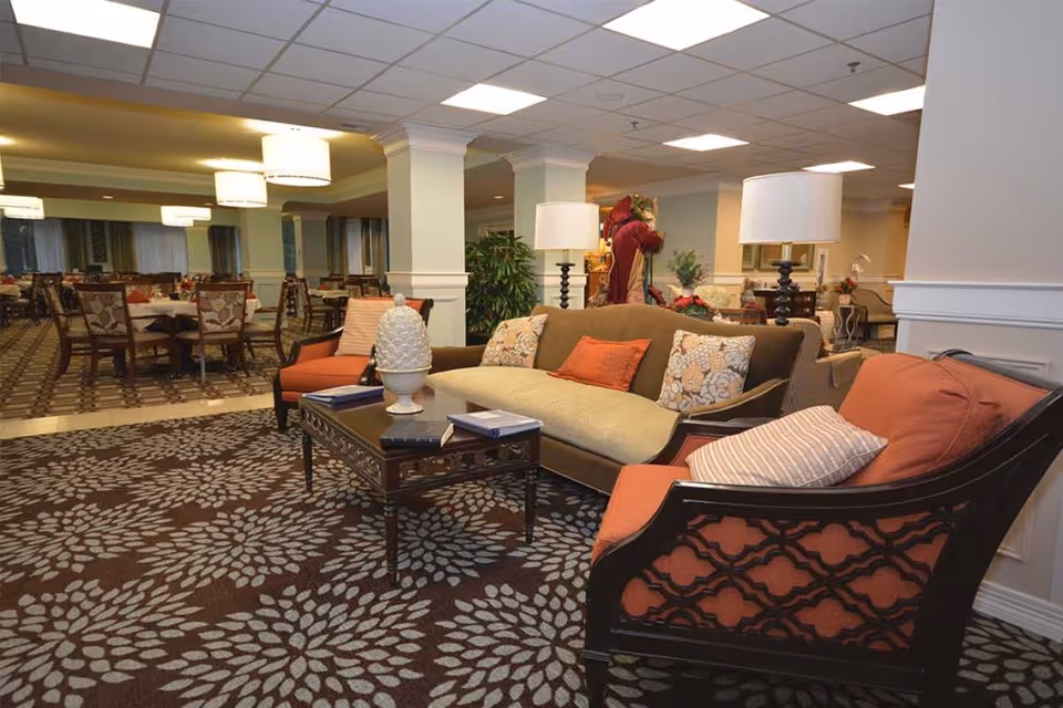 A cozy and well-lit common area in a retirement community featuring a seating arrangement with a sofa and two armchairs around a coffee table. The space has patterned carpeting, decorative pillows, table lamps, and a dining area with multiple tables and chairs in the background.