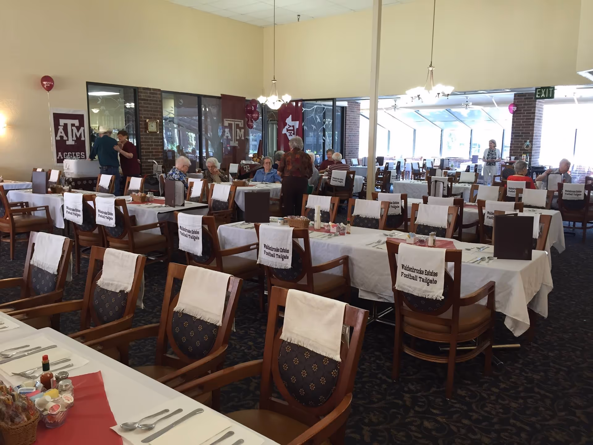 Dining room with tables and chairs set for a themed tailgate event, several residents seated and Texas A&M banners on the walls.