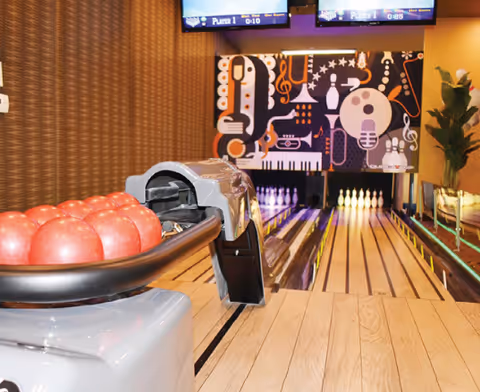 Indoor bowling alley with a rack of red bowling balls in the foreground and two lanes extending towards a colorful wall decorated with musical instrument graphics and pins set up at the end.