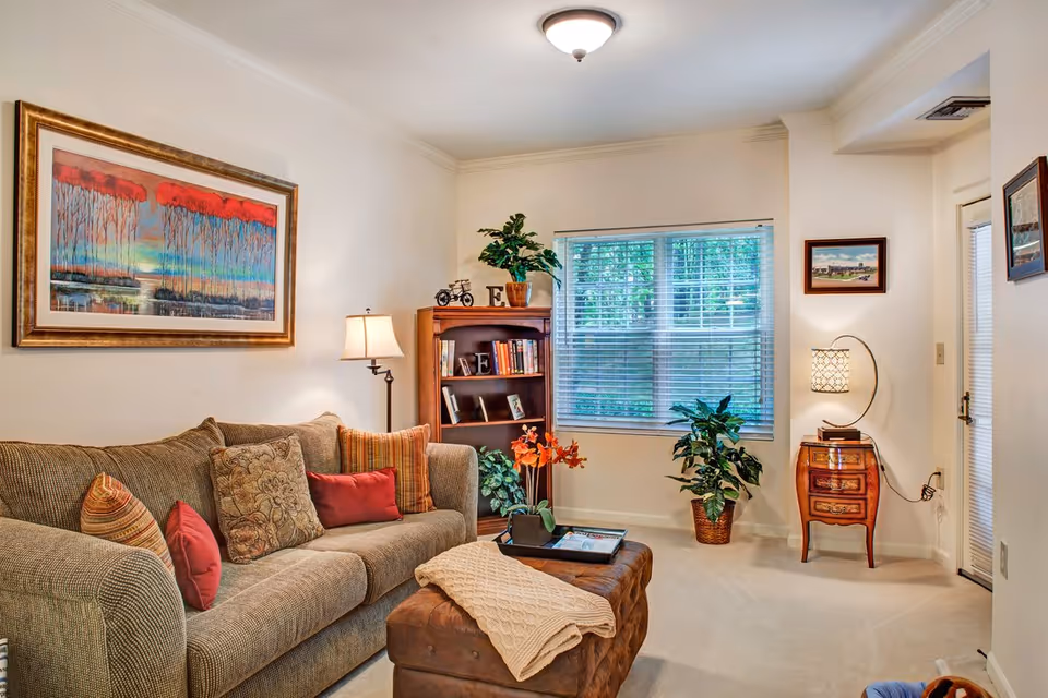 Cozy living room with a sofa, ottoman, bookshelf, potted plants, and a window with blinds.