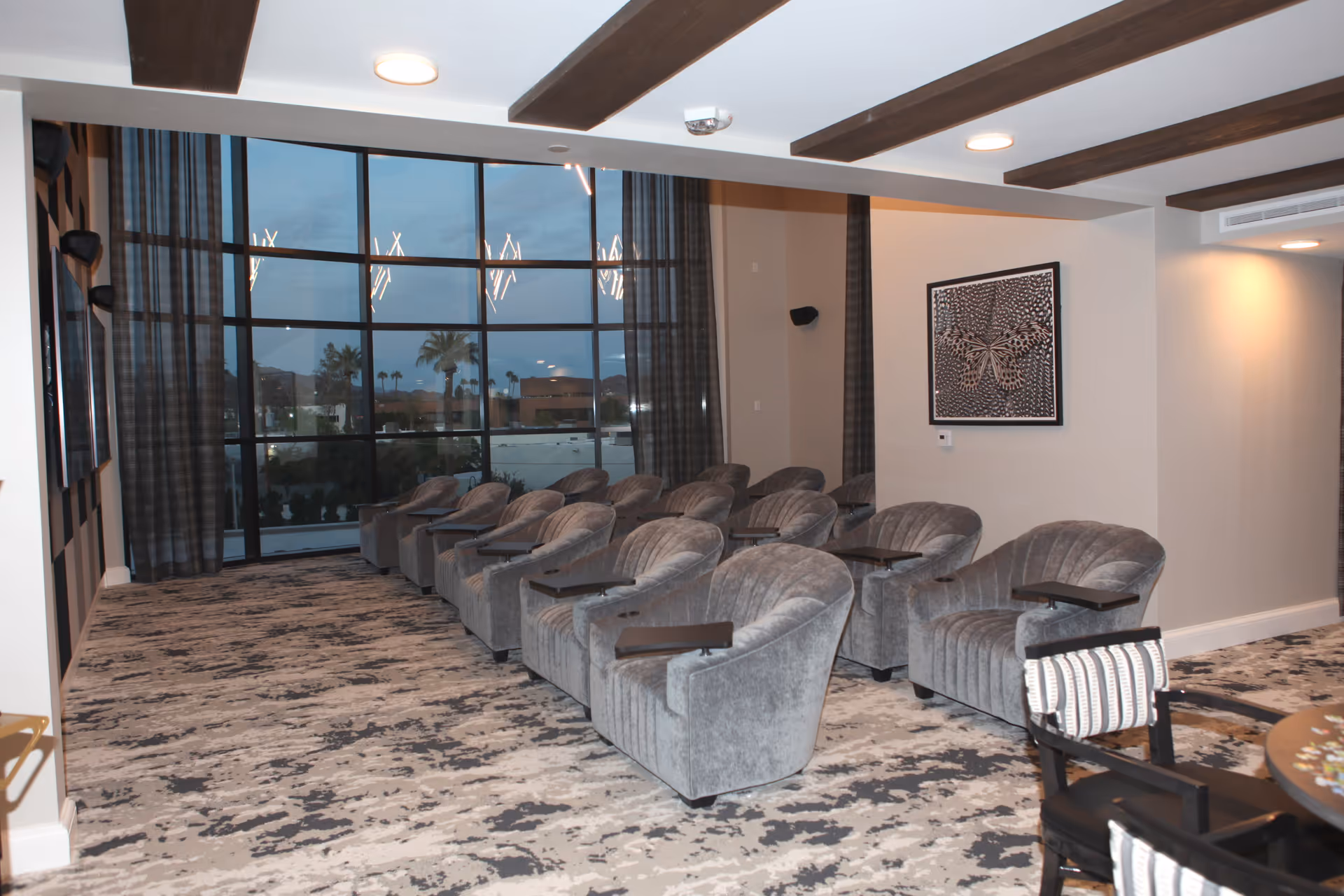 A modern lounge or media room with two rows of gray upholstered armchairs, each equipped with a small black tray table. The room features a large floor-to-ceiling window with a view of palm trees and buildings outside. The ceiling has exposed wooden beams and recessed lighting. A framed artwork of a butterfly hangs on the beige wall to the right.
