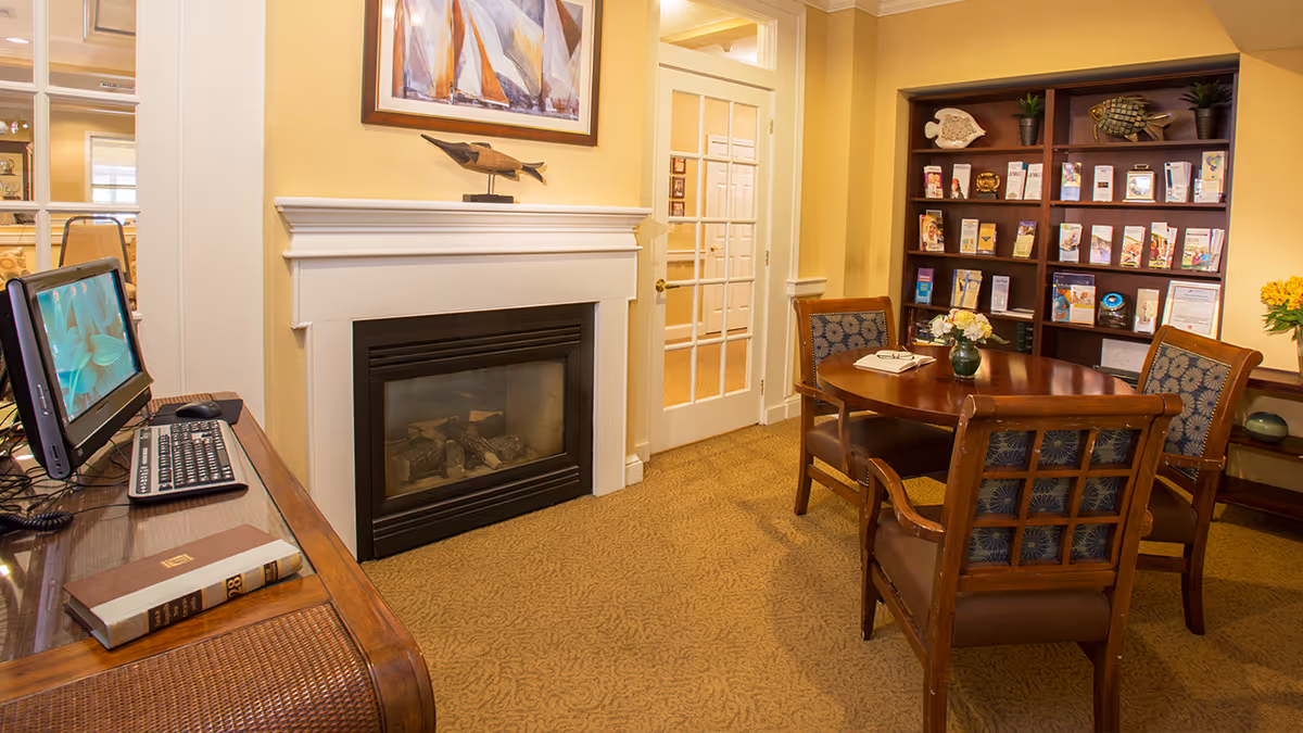 A cozy interior room with a fireplace, a wooden table surrounded by four cushioned chairs, a bookshelf filled with brochures and decorative items, and a computer workstation on a wooden desk. The walls are painted yellow and there is a glass-paneled door in the background.