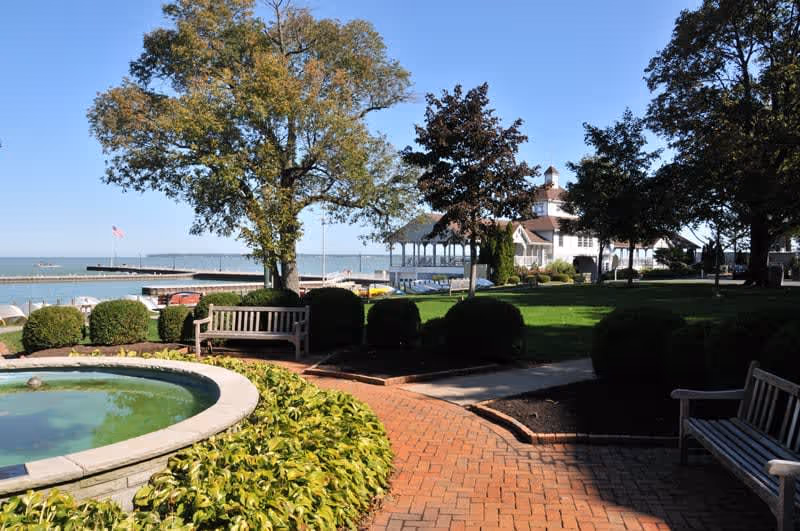A peaceful outdoor garden area with a circular water feature surrounded by green plants, wooden benches, trimmed bushes, and large trees. In the background, there is a white building with a covered porch near a body of water under a clear blue sky.