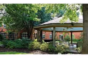 Brick senior living building with a covered gazebo and outdoor seating surrounded by trees and landscaping.