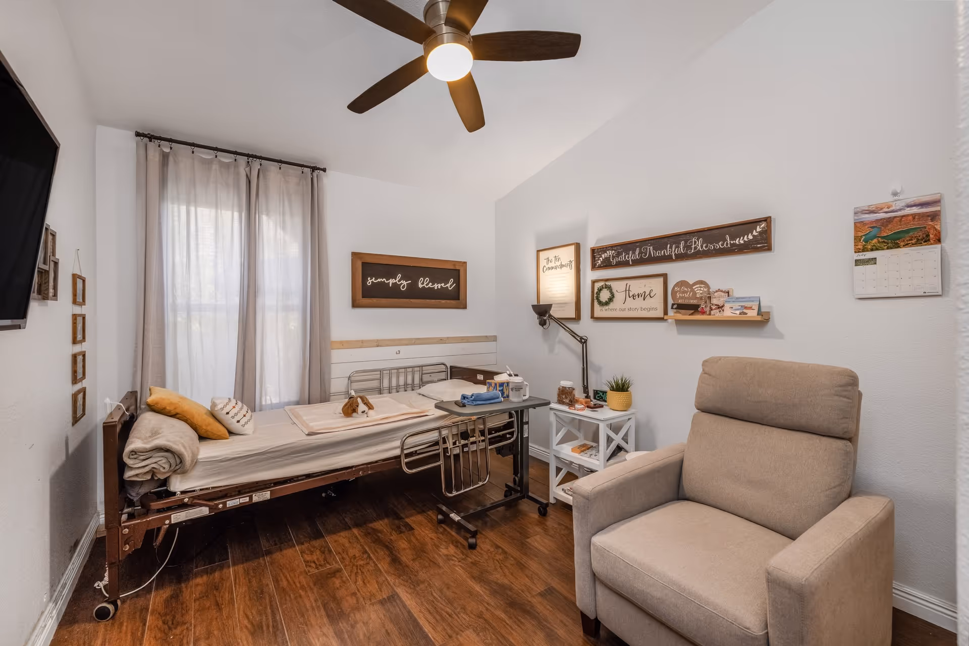 A cozy assisted living bedroom with a hospital-style bed, beige armchair, wooden floor, and a ceiling fan. The bed is made with light-colored bedding and has a small stuffed animal on it. The walls are decorated with framed inspirational quotes and a calendar. A small white side table holds a lamp, plant, and other small items.