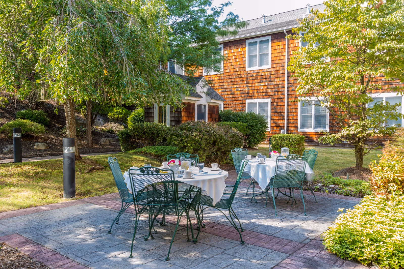 Outdoor patio area with two round tables covered with white tablecloths, set with cups, glasses, and a pitcher. Green metal chairs surround the tables. The patio is paved with stone tiles and bordered by bushes and trees. In the background, there is a building with brown shingle siding and white-framed windows.