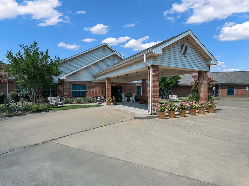 Exterior view of Chapters Living of Maumelle showing a covered entrance with brick pillars, potted flowers, benches, and a clear blue sky with scattered clouds.