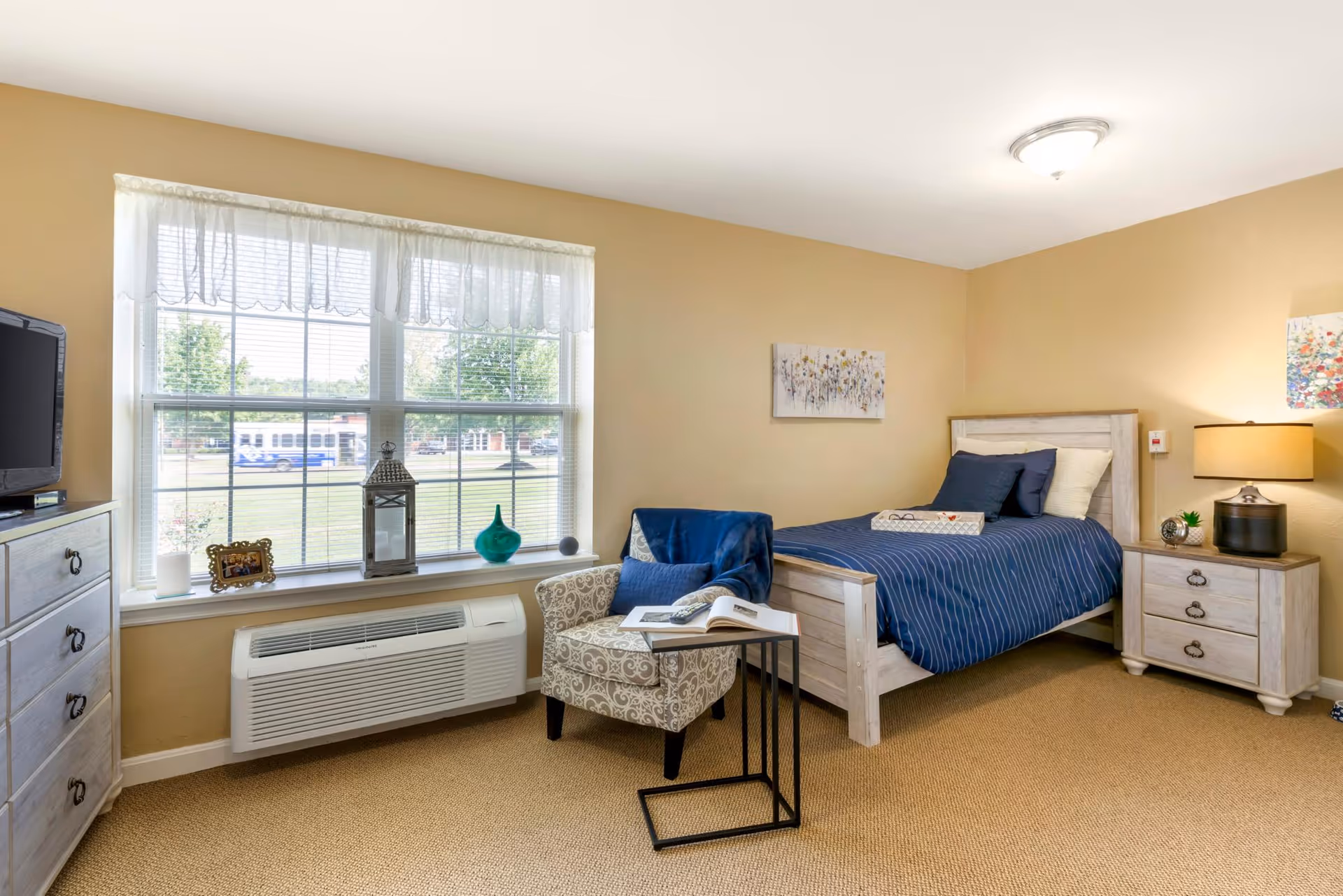 Bright furnished bedroom with a single bed, nightstand, dresser and armchair beside a large window.