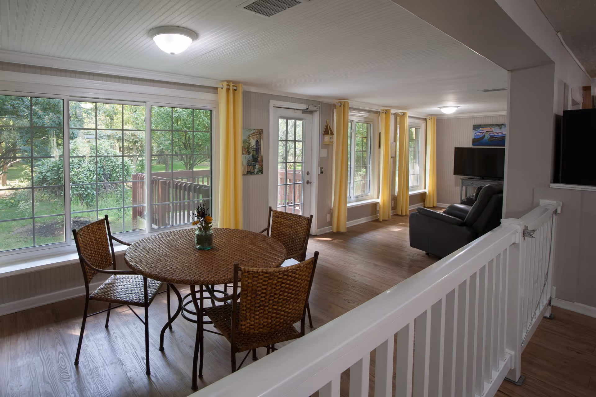 Interior view of a senior living facility room with large windows and yellow curtains letting in natural light. There is a round wicker table with four matching chairs near the windows. The room extends into a living area with a black leather sofa and a flat-screen TV mounted on the wall. The floor is wood, and there is a white railing in the foreground.