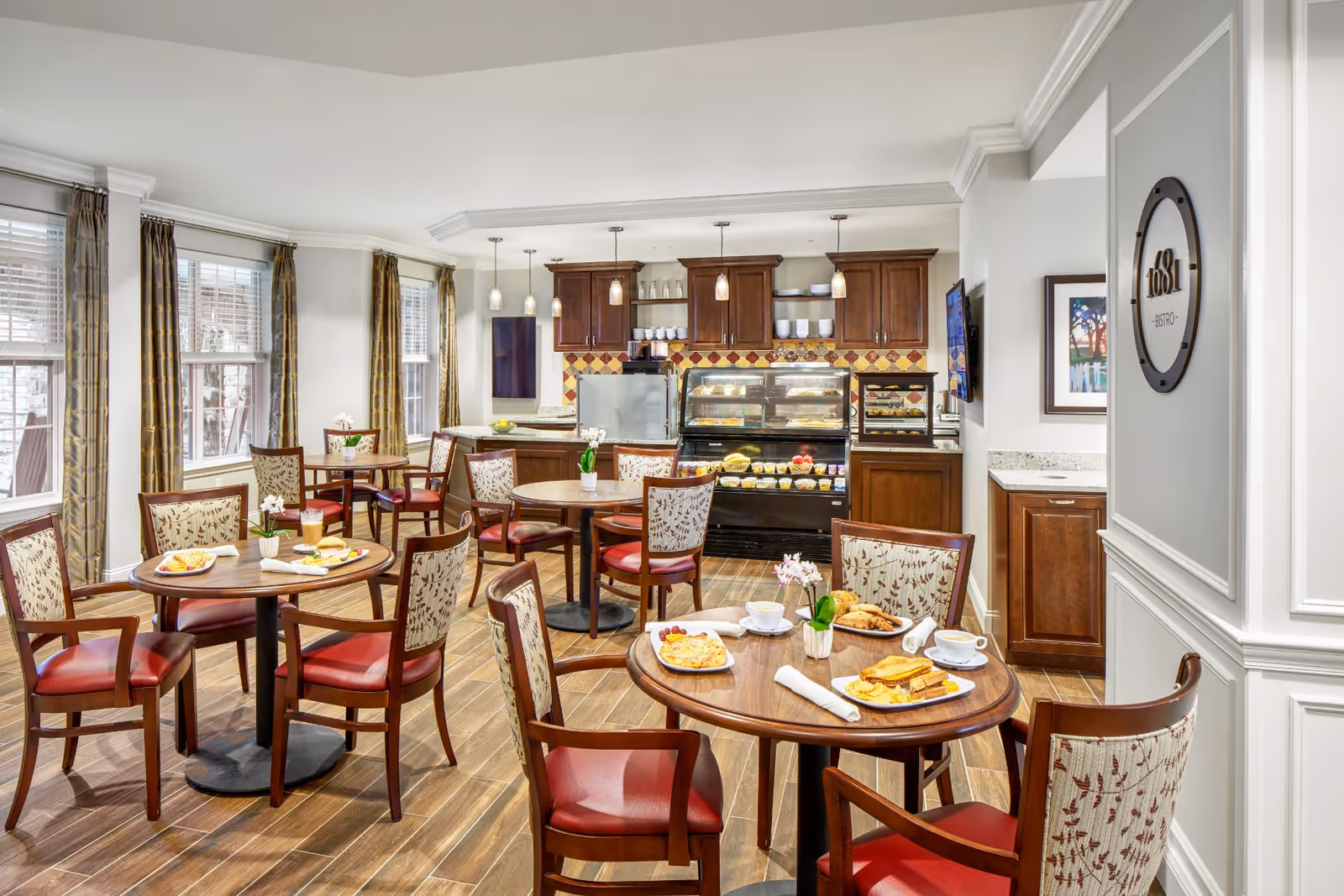 Bright bistro-style dining room with round wooden tables, red-upholstered chairs, place settings, and a food display counter in the back.