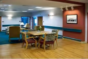 Interior view of a nursing and rehab center common area with wooden flooring, a table surrounded by four chairs with floral upholstery, a seating area with armchairs and a sofa, a lamp, and framed artwork on the walls.