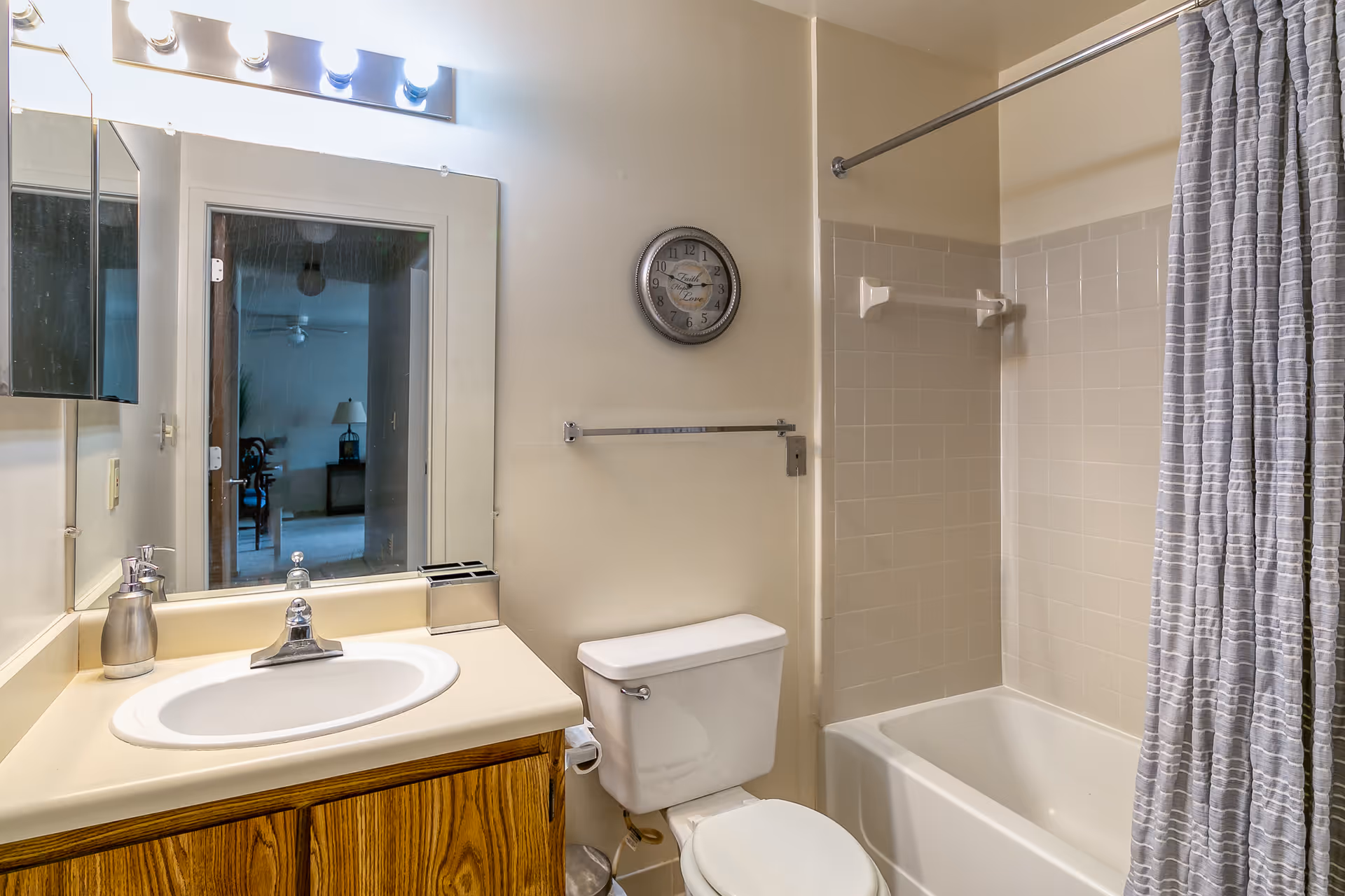 A bathroom with a white sink and wooden cabinet, a toilet, a bathtub with a gray and white striped shower curtain, a wall-mounted clock, and a large mirror reflecting an adjacent room.