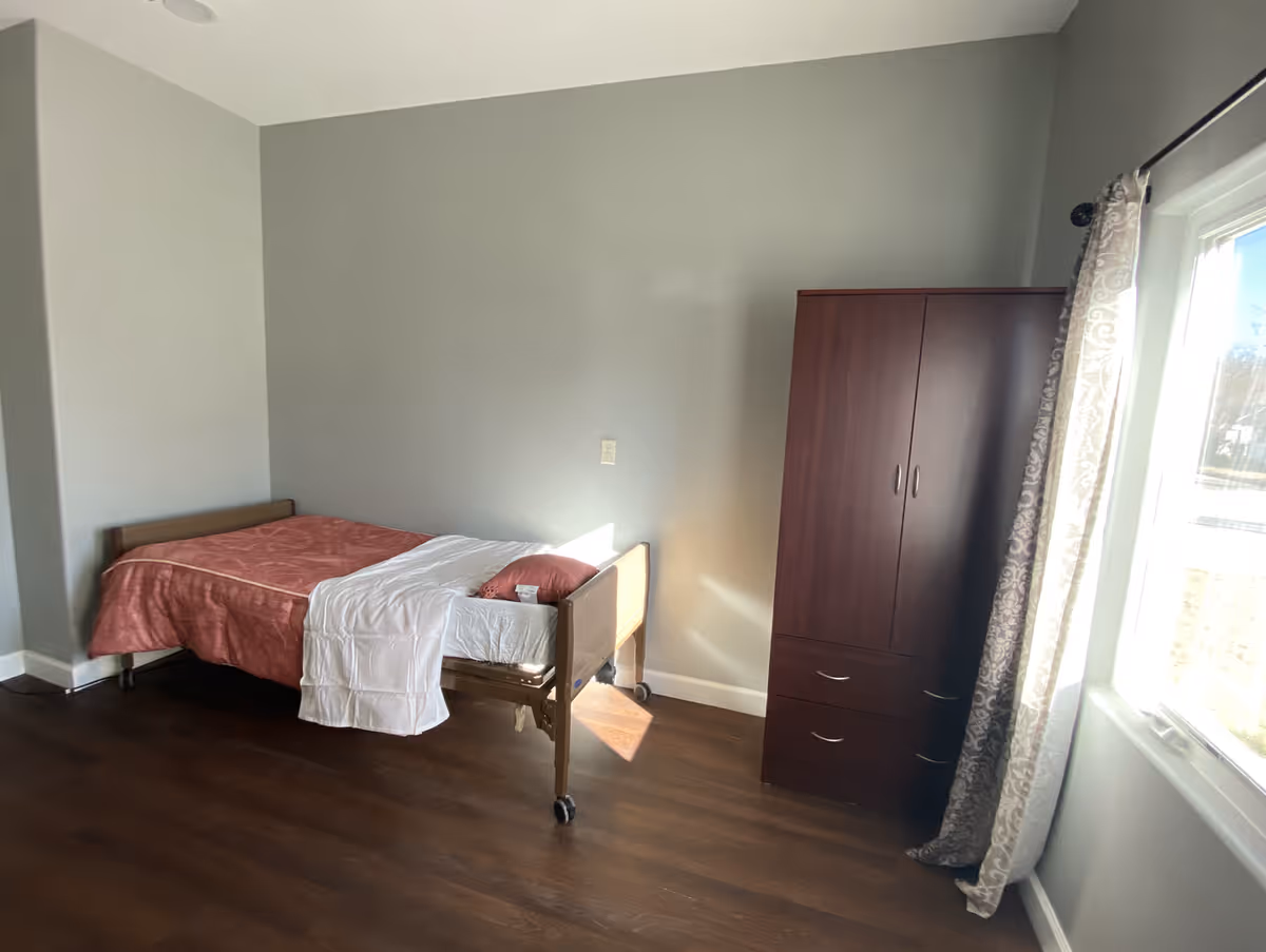 A simple bedroom with a single bed covered with a red blanket and white sheet, a wooden wardrobe, and a window with patterned curtains letting in natural light. The room has light gray walls and dark wooden flooring.