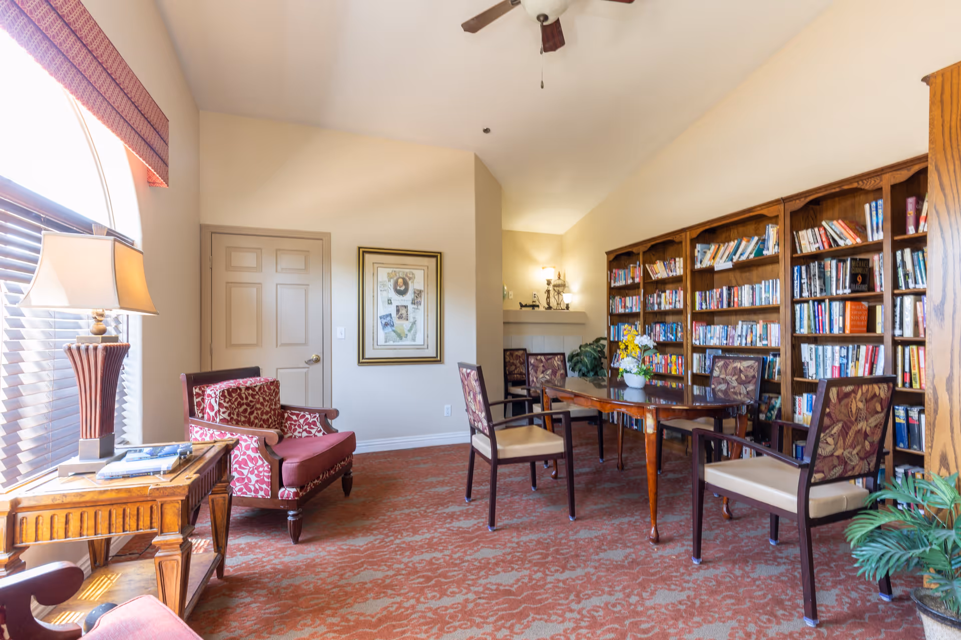A cozy reading room with a large wooden bookshelf filled with books along one wall, a wooden table surrounded by four chairs with floral upholstery, a red patterned armchair, a wooden side table with a lamp, and a framed picture on the wall. The room has a large window with blinds and a ceiling fan.