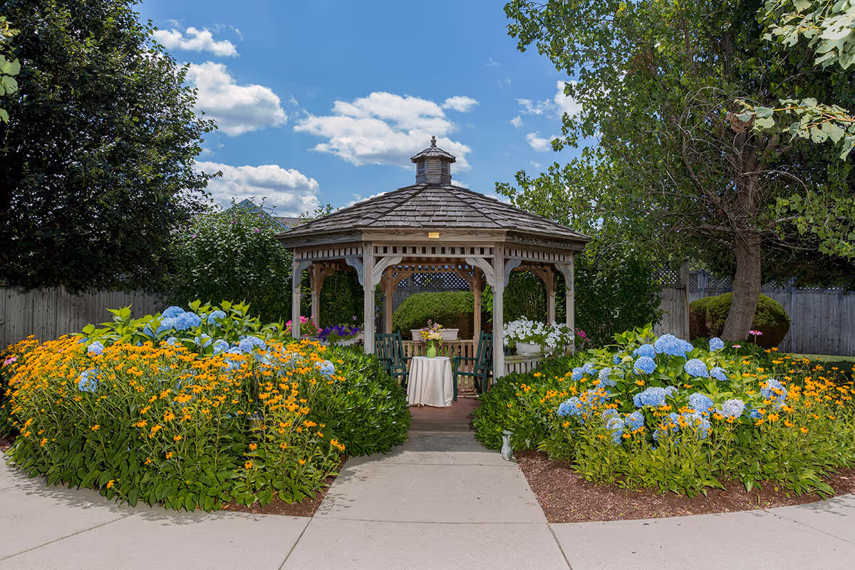A wooden gazebo surrounded by vibrant flower beds with yellow and blue flowers, set in a garden area with trees and a clear blue sky with some clouds.