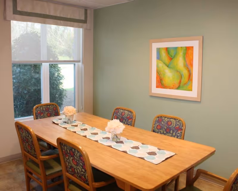 Small dining room with a rectangular wooden table, six upholstered chairs, a decorative runner with floral centerpieces, a window showing greenery, and a framed artwork on a green wall.