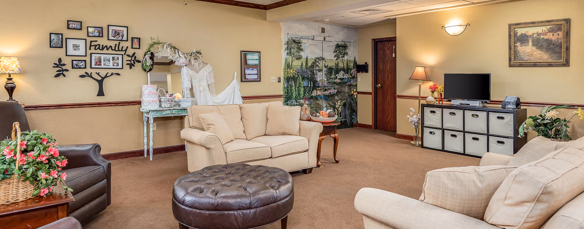 A cozy living room in a senior living facility with beige walls and carpet. The room features two beige sofas, a dark brown leather armchair, and a round tufted ottoman in the center. On the left wall, there is a decorative family photo arrangement with the word 'Family' and a small table with a lamp and flowers. The back wall has a painted mural of a scenic outdoor landscape, a wooden door, and a small table with a lamp and TV on a storage unit with fabric bins. There are framed pictures and plants adding to the warm, homey atmosphere.