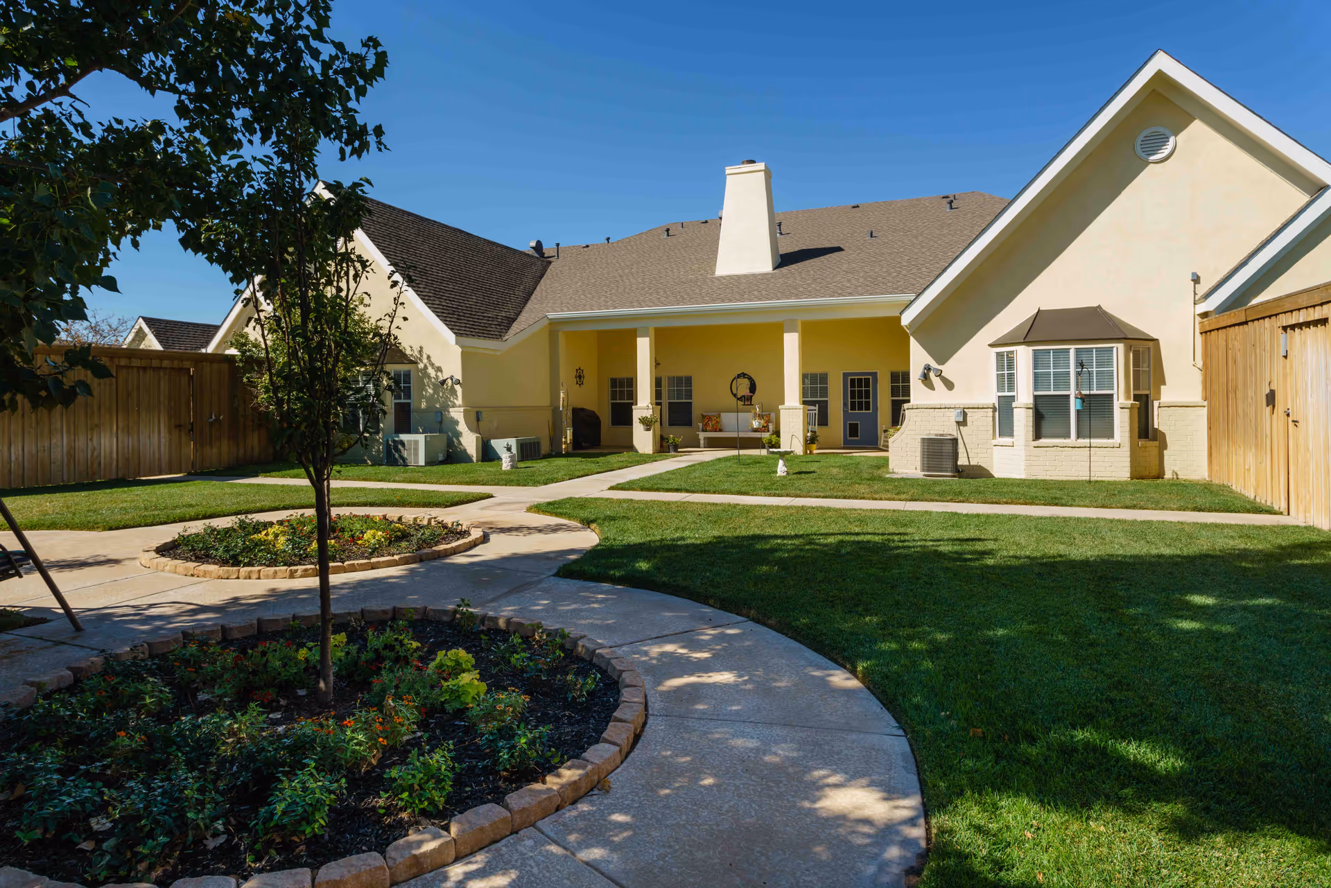 Outdoor courtyard area of a senior living facility with a circular flower bed, green grass, paved walkways, and a yellow building with a covered porch and multiple windows under a clear blue sky.