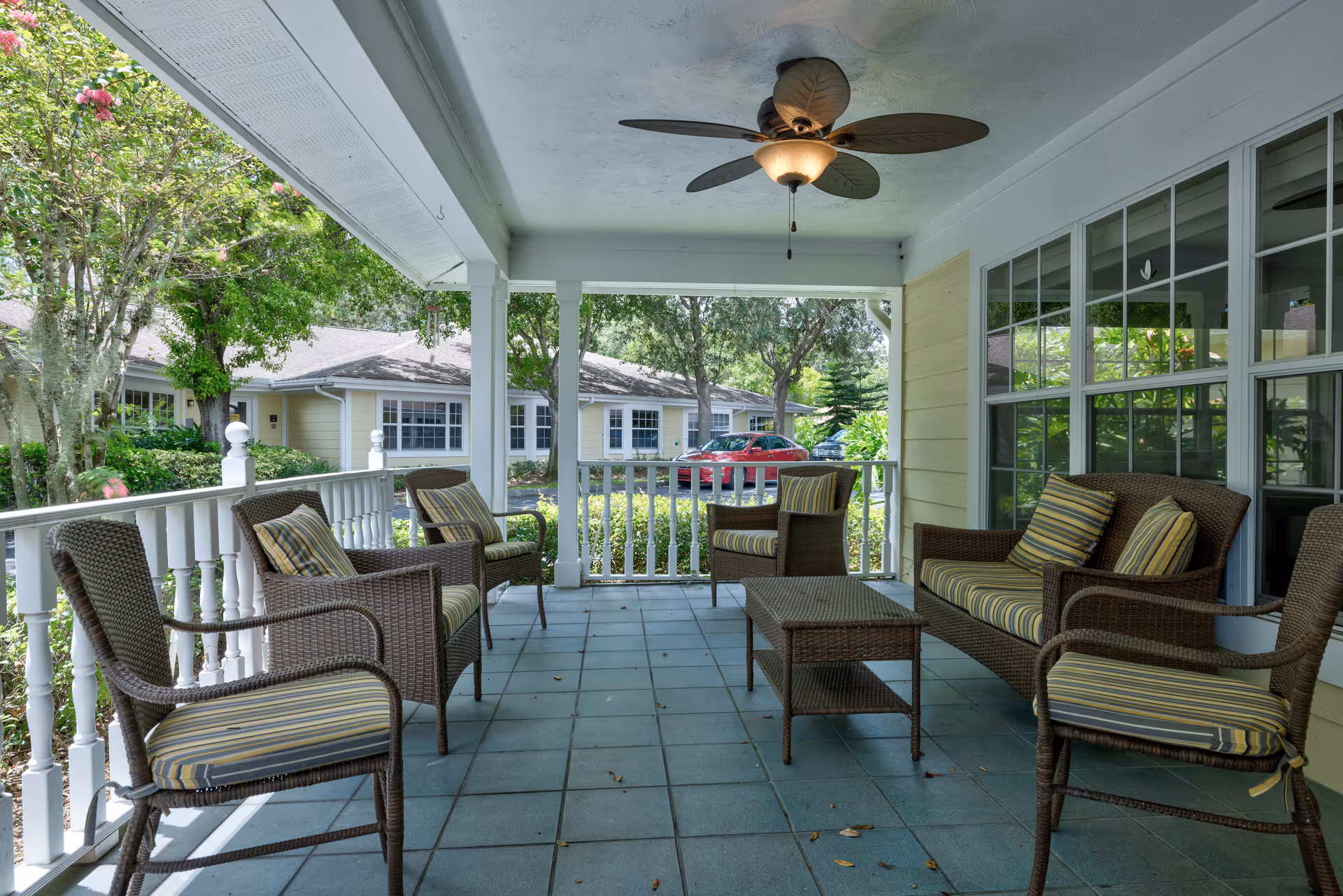 Covered outdoor patio area with wicker furniture including chairs and a loveseat with striped cushions, a coffee table, ceiling fan, and a view of trees, bushes, and a red car parked outside.