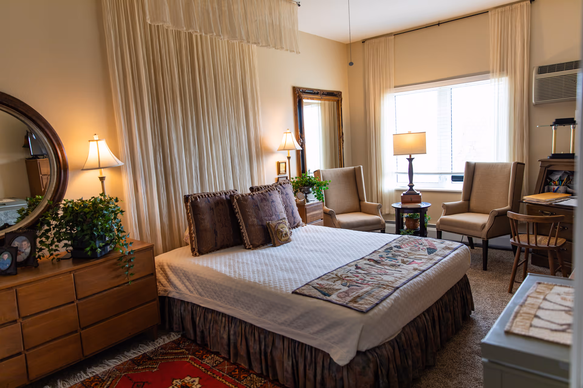A cozy bedroom with a large bed covered in a white quilt and decorative pillows. There is a wooden dresser with a round mirror and potted plant on the left side. Two beige armchairs and a small round table with a lamp are positioned near a window with sheer curtains. The room has warm lighting and a carpeted floor with a patterned rug.