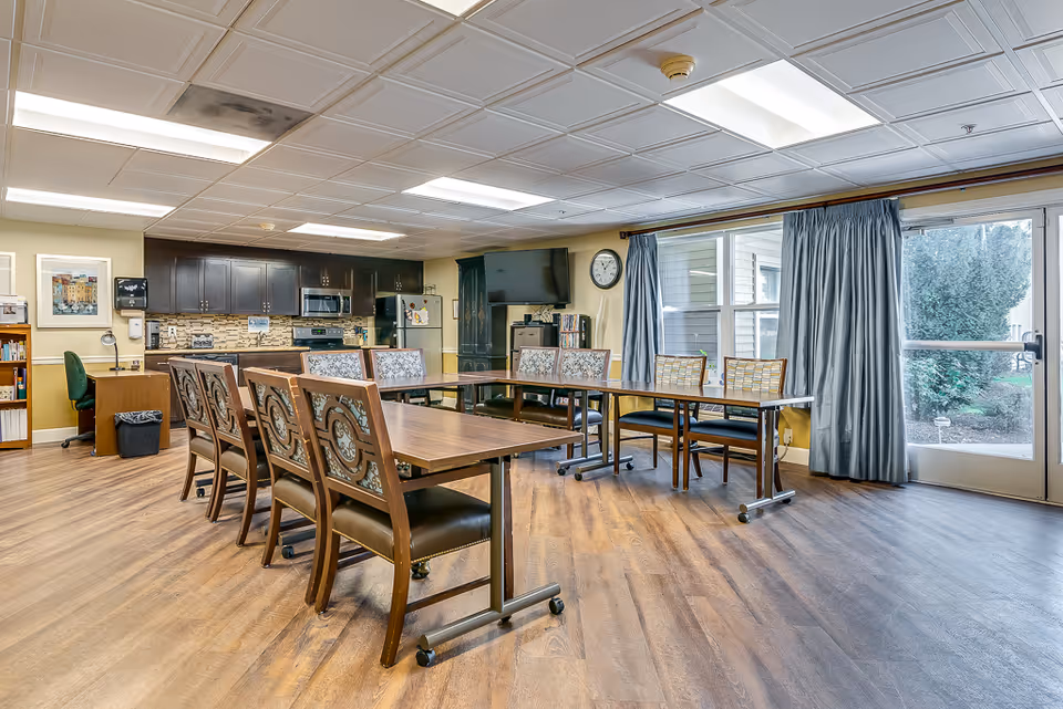 A bright senior living common room with tables and chairs, a kitchenette and TV, and a glass door leading outside.