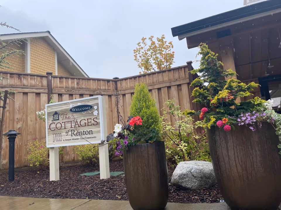 Entrance sign for The Cottages of Renton beside large planters with flowers and a wooden fence backdrop.