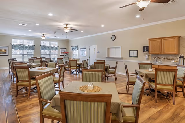 A spacious dining room with multiple wooden tables covered with beige tablecloths and surrounded by green cushioned chairs. The room has wooden flooring, ceiling fans with lights, large windows with blue valances, and a small serving window in the wall. There are cabinets and a coffee station on the right side of the room.