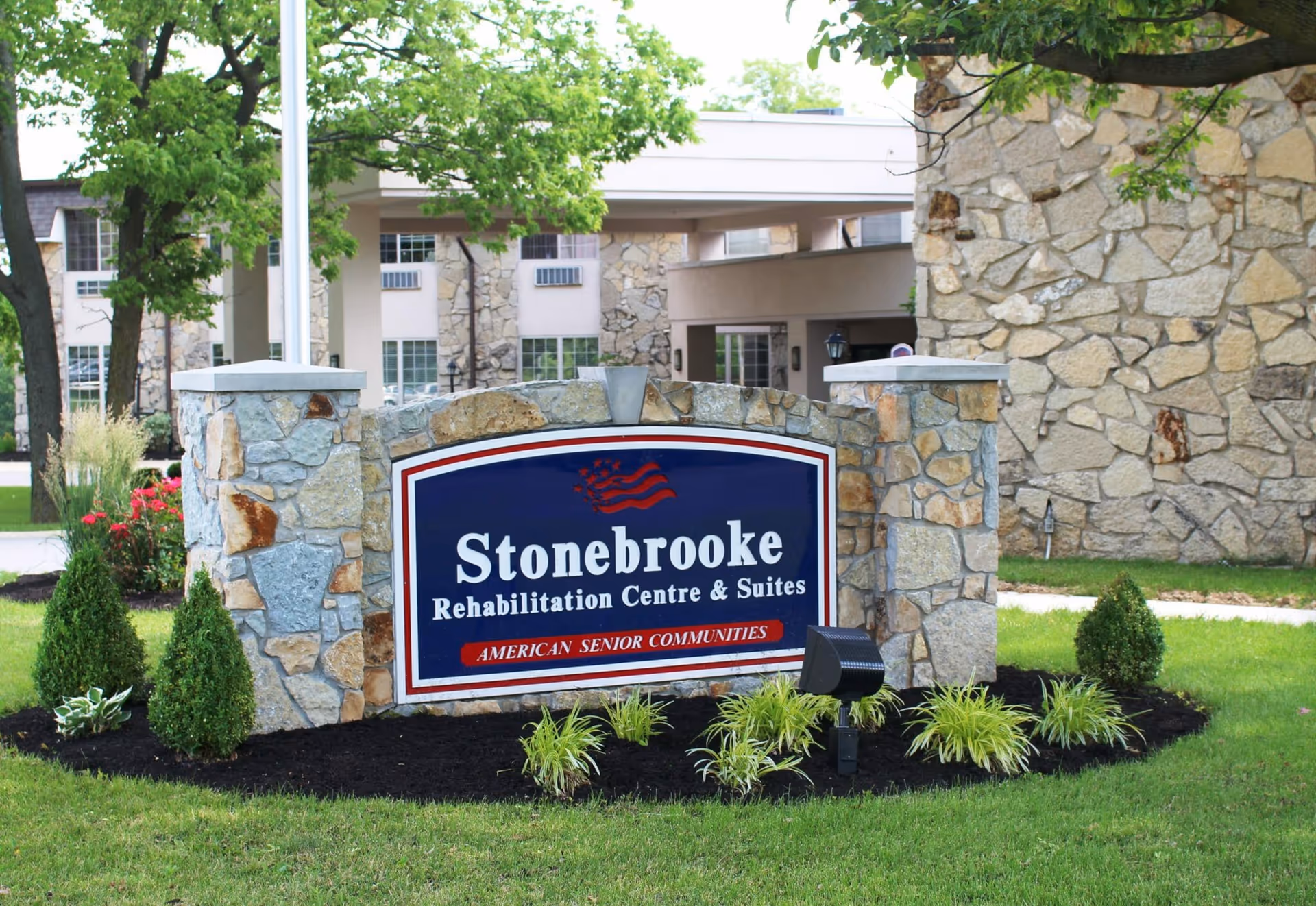 Stonebrooke Rehabilitation Centre & Suites sign made of stone with a blue background and white text, surrounded by greenery and plants, with part of the building visible in the background.