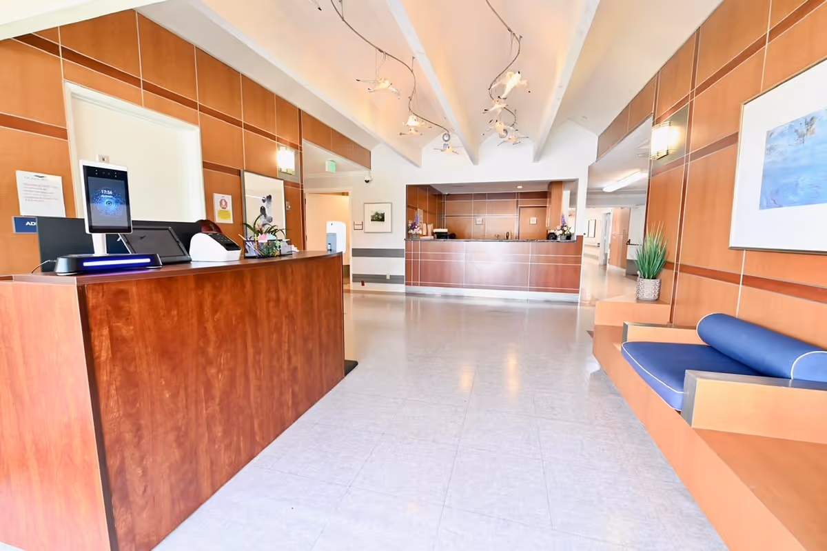 Bright and clean reception area of Ocean Pointe Healthcare Center featuring a wooden front desk with a digital check-in device, a seating area with blue cushioned bench, potted plants, and modern lighting fixtures on the ceiling.