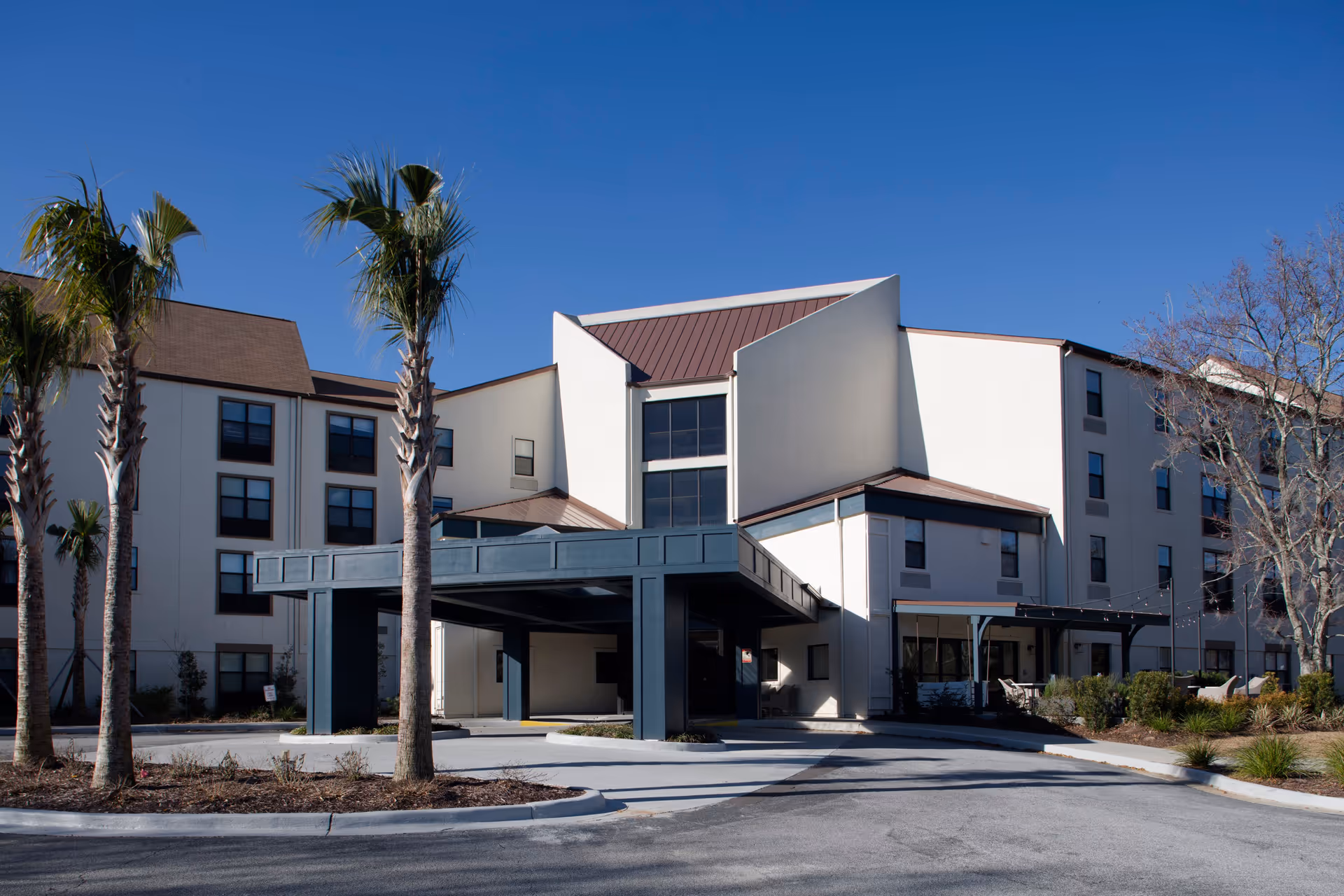 Exterior view of a multi-story senior living facility building with a covered entrance, palm trees, and clear blue sky.
