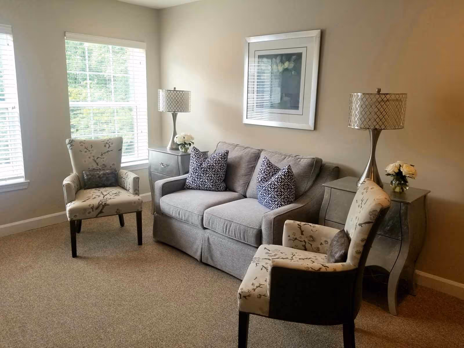 Cozy living room with a gray loveseat, two patterned armchairs, side tables with lamps, and a framed picture by bright windows.