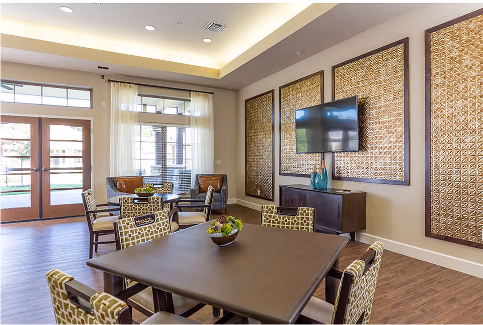 A bright and modern common area with a large square wooden table surrounded by patterned chairs. In the background, there are two armchairs with brown cushions near large windows and glass doors letting in natural light. On the wall, there are three large decorative panels with a geometric pattern and a flat-screen TV mounted above a dark wooden cabinet with decorative vases.