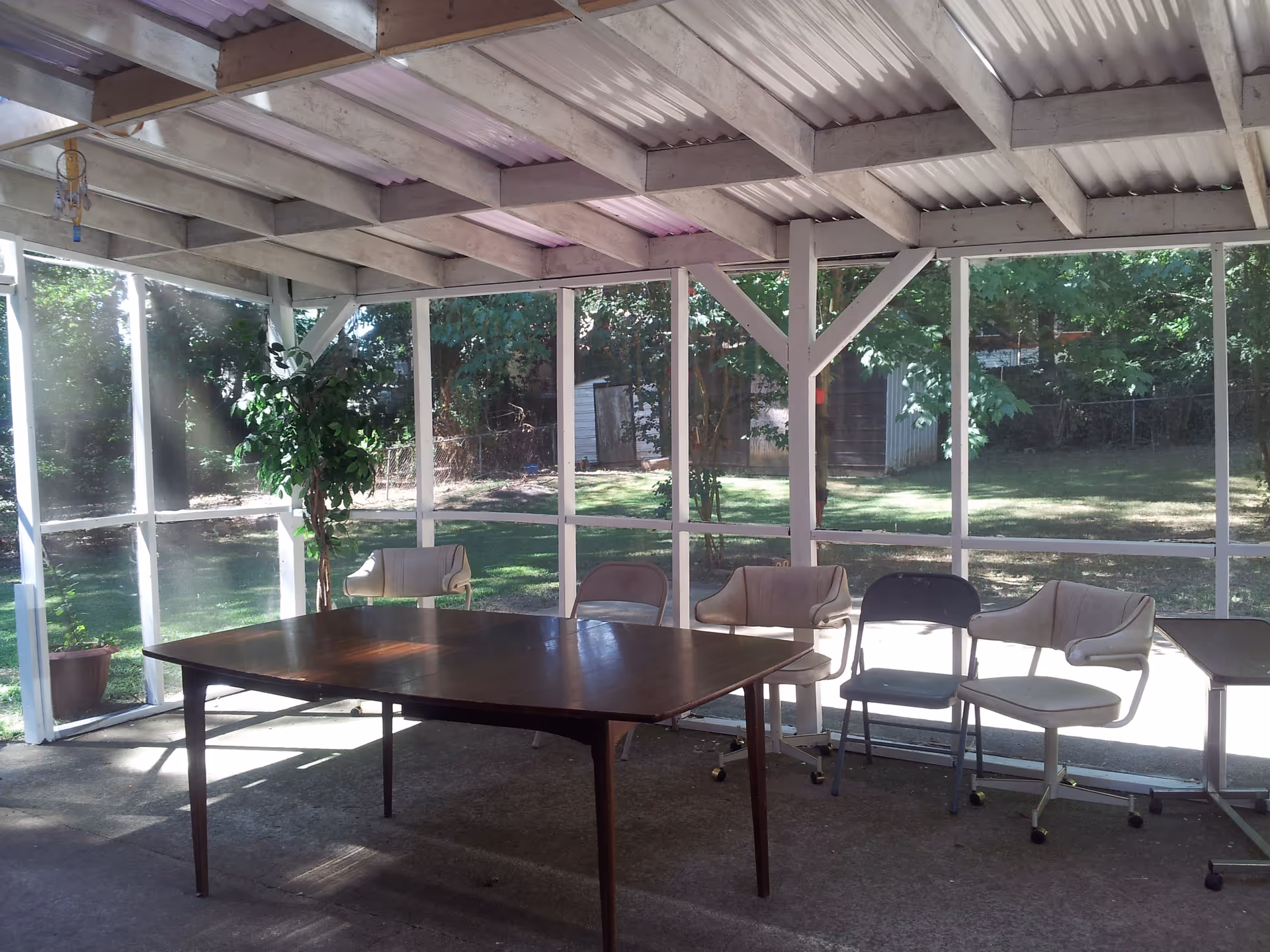 A screened-in porch area with a wooden table and several chairs arranged around it. The porch has a corrugated roof and large screened windows that provide a view of a grassy backyard with trees and a shed.