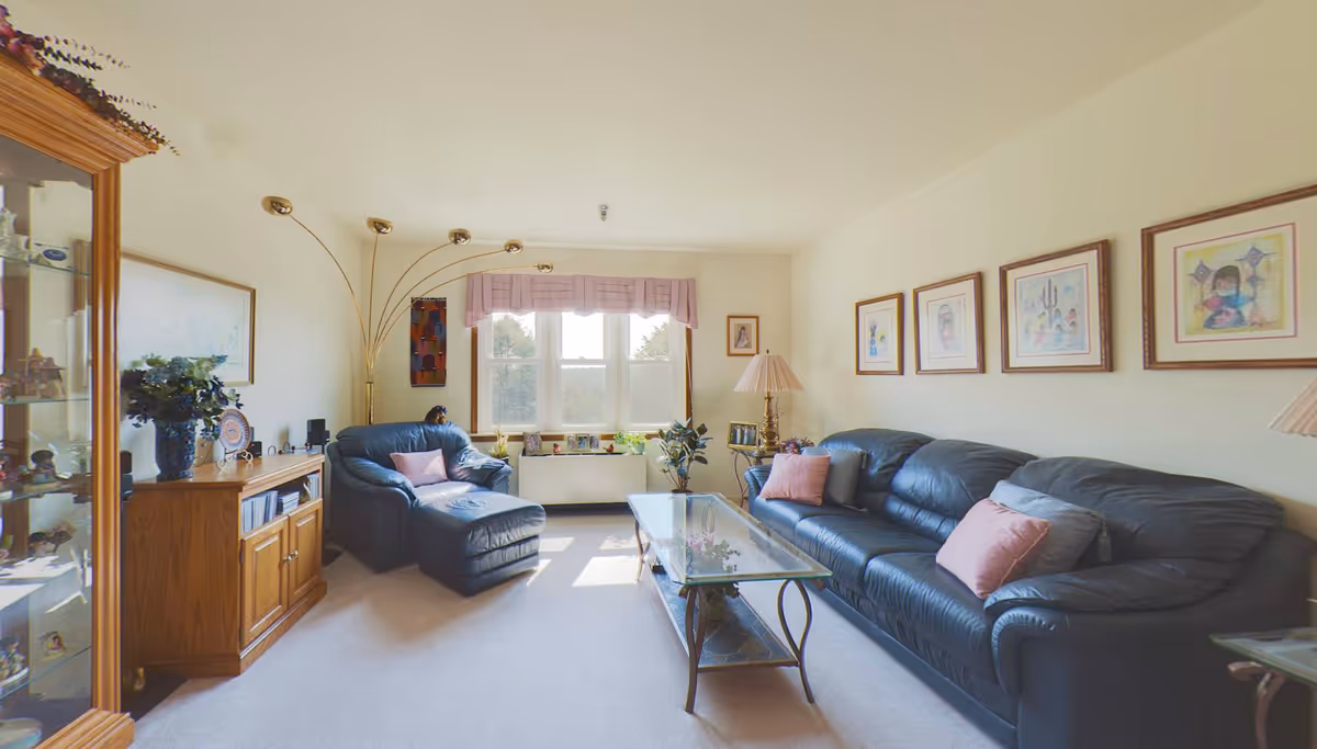 A cozy living room with a large window letting in natural light. The room features a dark blue leather sofa and matching armchair with pink and gray cushions. A glass coffee table with a metal frame sits in the center. On the left side, there is a wooden cabinet with decorative items and a tall, curved floor lamp with multiple lights. Several framed artworks hang on the walls, and there are plants and photo frames on the window sill and side table.