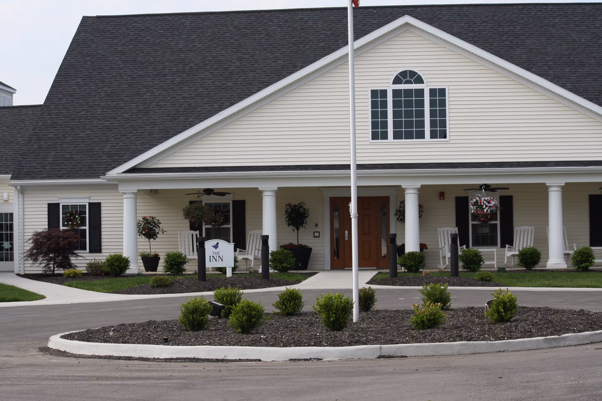 Front exterior view of a senior living community building named The Inn, featuring a beige facade with white columns, a dark shingled roof, a central entrance with double wooden doors, rocking chairs on the porch, hanging flower baskets, and landscaped bushes in front.