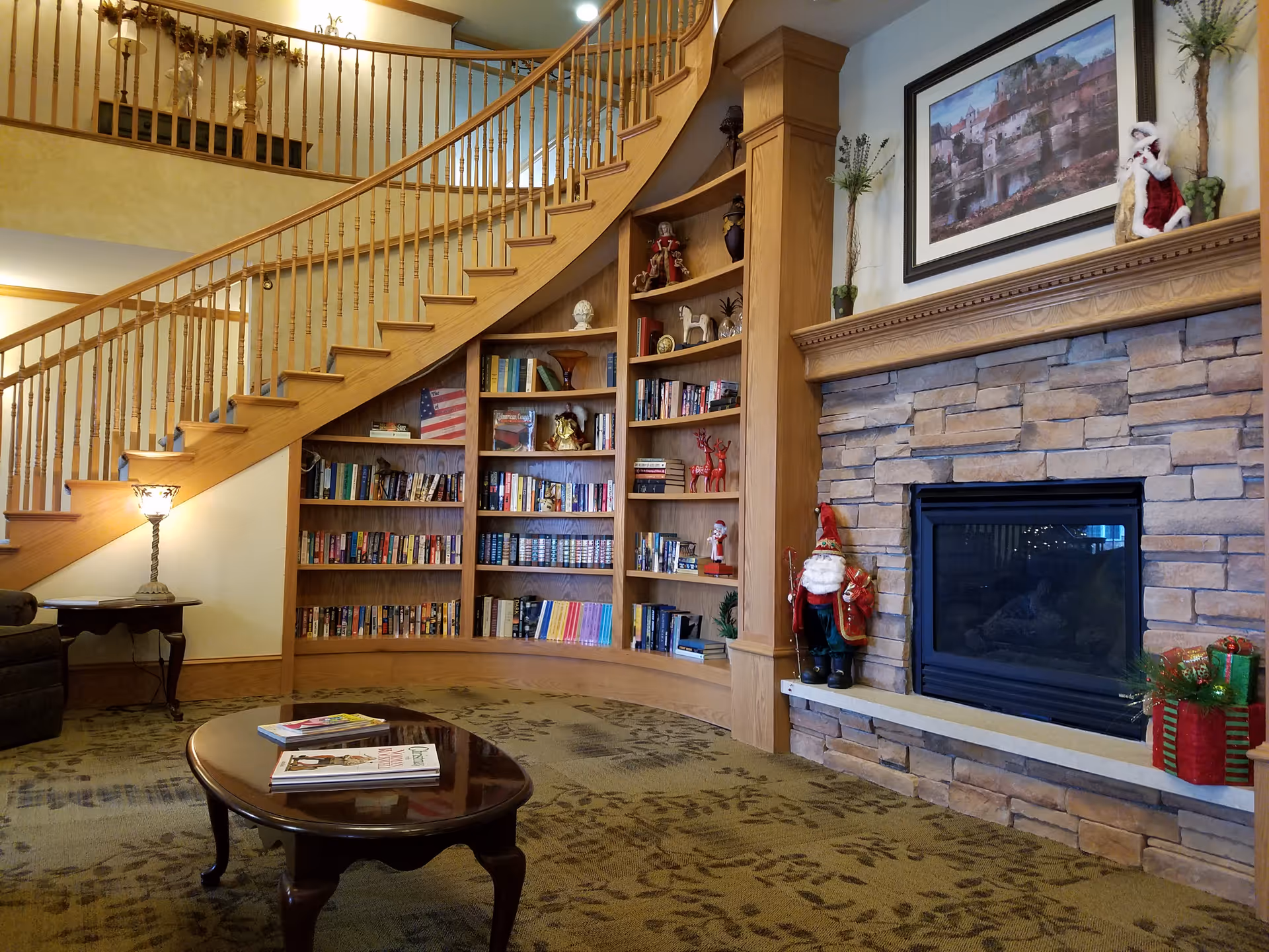 Cozy living room area with a curved wooden staircase and built-in bookshelves filled with books and decorative items. A stone fireplace with a wooden mantle is decorated with holiday figurines and a framed picture above it. A wooden coffee table with magazines sits on a patterned carpet, and a small side table with a lamp is next to a dark armchair.