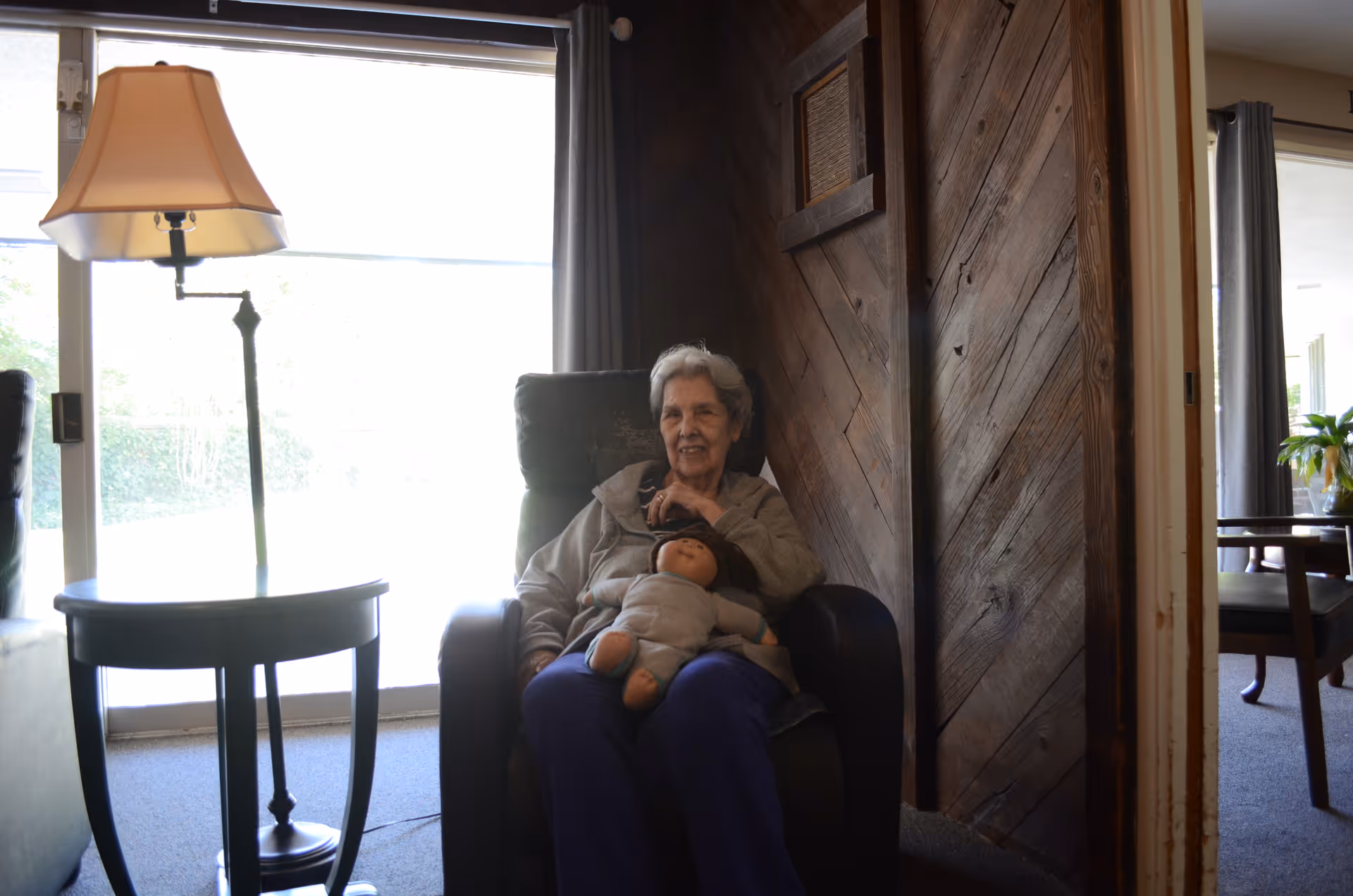 An elderly woman sitting in a dark armchair holding a doll, next to a round side table with a lamp. The room has wooden paneling on one wall and large windows with curtains letting in natural light. Another room with chairs and a table is visible through an open doorway.