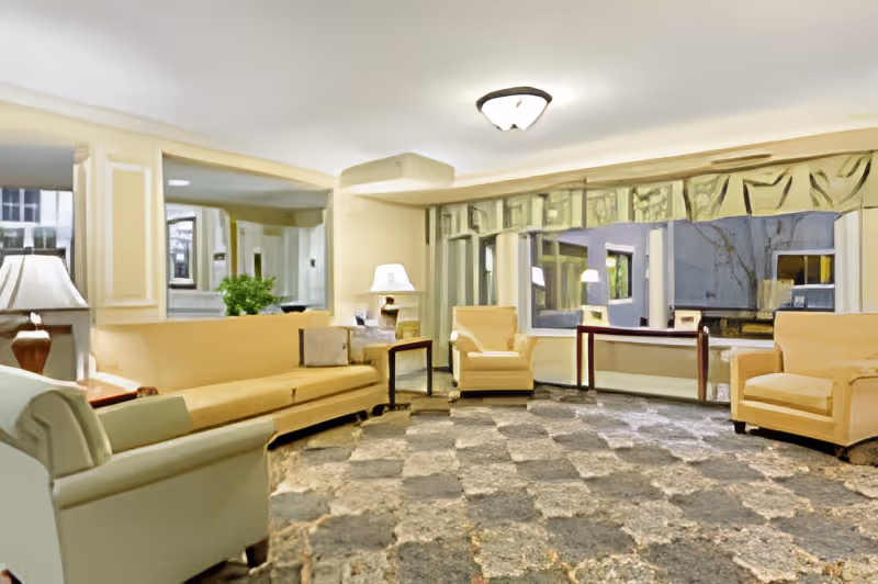 A cozy living room area with beige and light green armchairs and a long beige sofa arranged around a patterned carpet. There are two side tables with lamps, a large window with drapes, and a mirror on the wall reflecting part of the room.