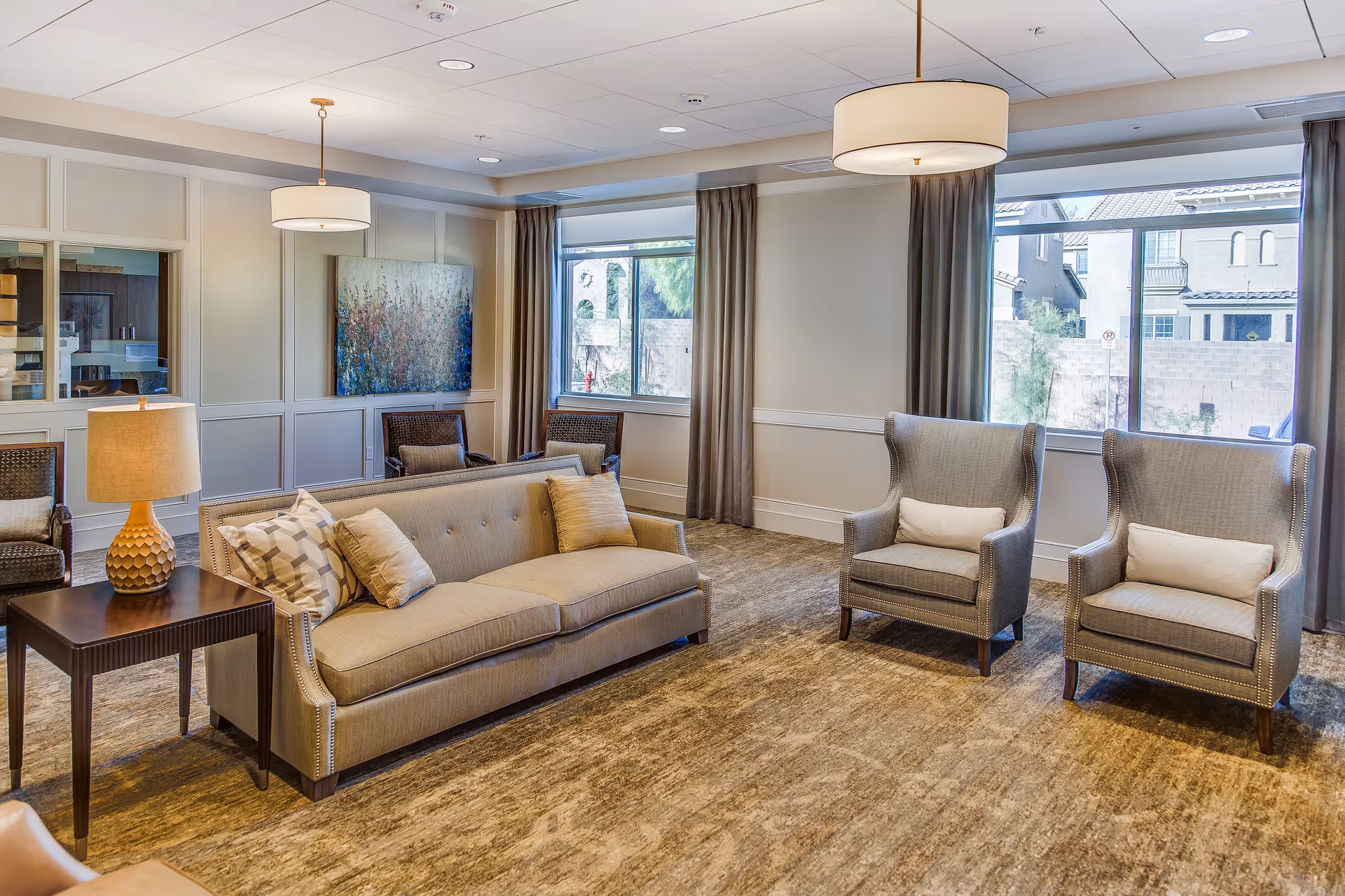 A well-lit living room with a beige sofa adorned with pillows, two gray armchairs with white cushions, a wooden side table with a lamp, and large windows with gray curtains letting in natural light. The room features a carpeted floor, wall paneling, and a large abstract painting on the wall.