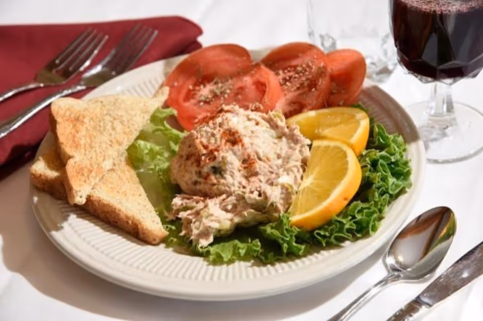 A plate with a serving of tuna salad on a bed of lettuce, garnished with lemon wedges and sliced tomatoes, accompanied by two triangular pieces of toasted bread. The plate is set on a white tablecloth with a spoon, knife, fork, a glass of water, and a glass of red wine nearby.