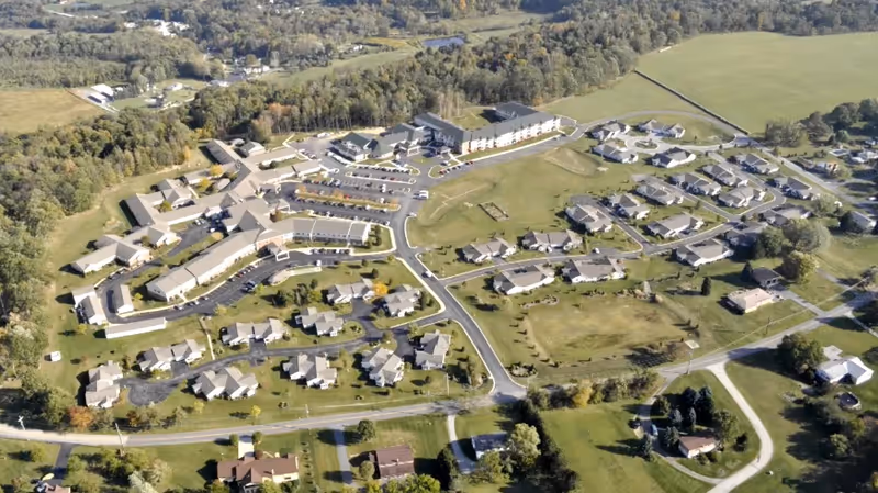 Aerial view of Laurel View Village senior living facility showing multiple residential buildings, parking lots, roads, and surrounding green fields and trees.