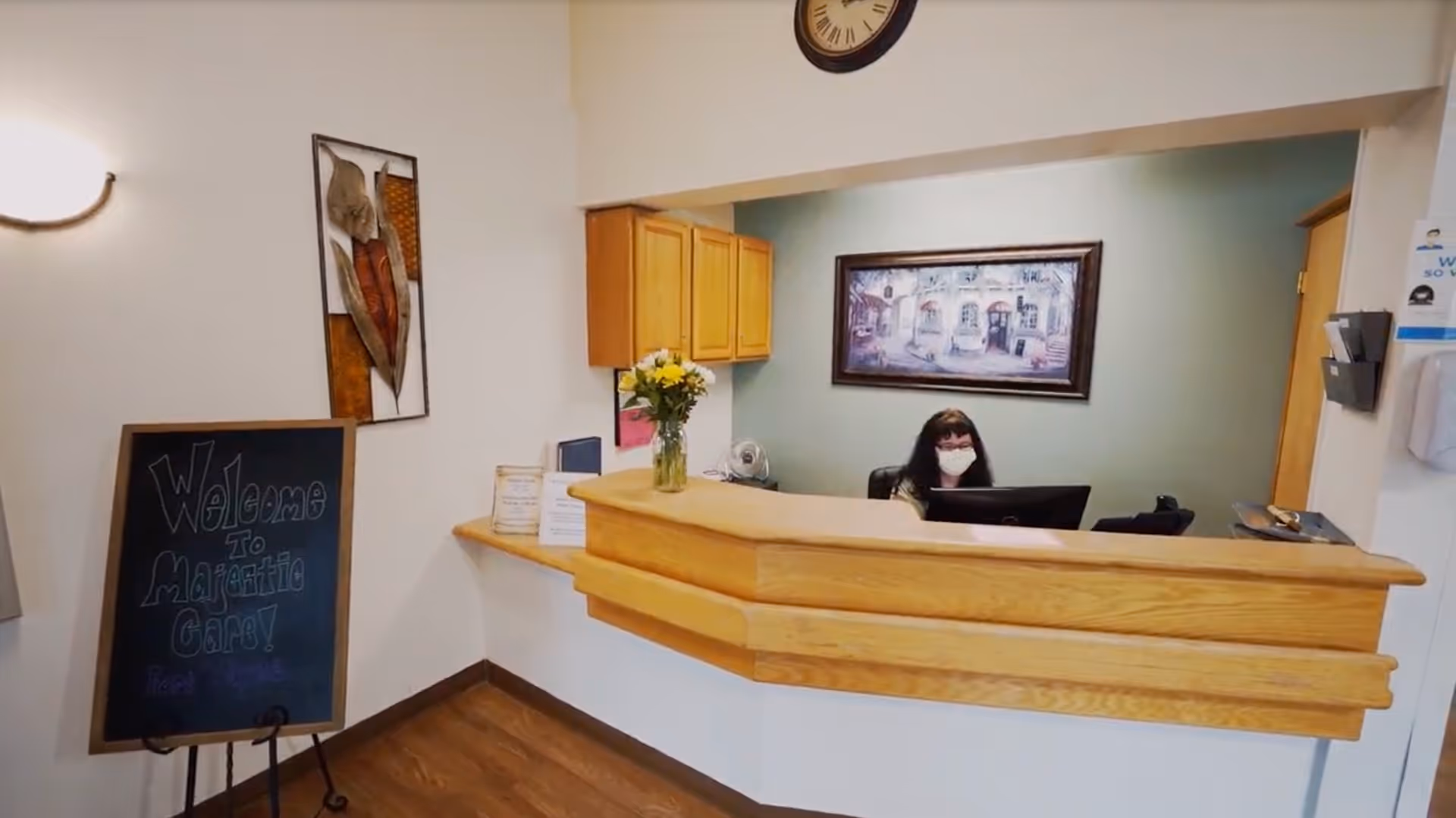 Reception area of Majestic Care of Fort Wayne with a wooden front desk, a woman wearing a face mask seated behind the desk, a wall clock above, a framed painting on the wall, a vase with flowers on the counter, and a chalkboard sign that says 'Welcome to Majestic Care!'