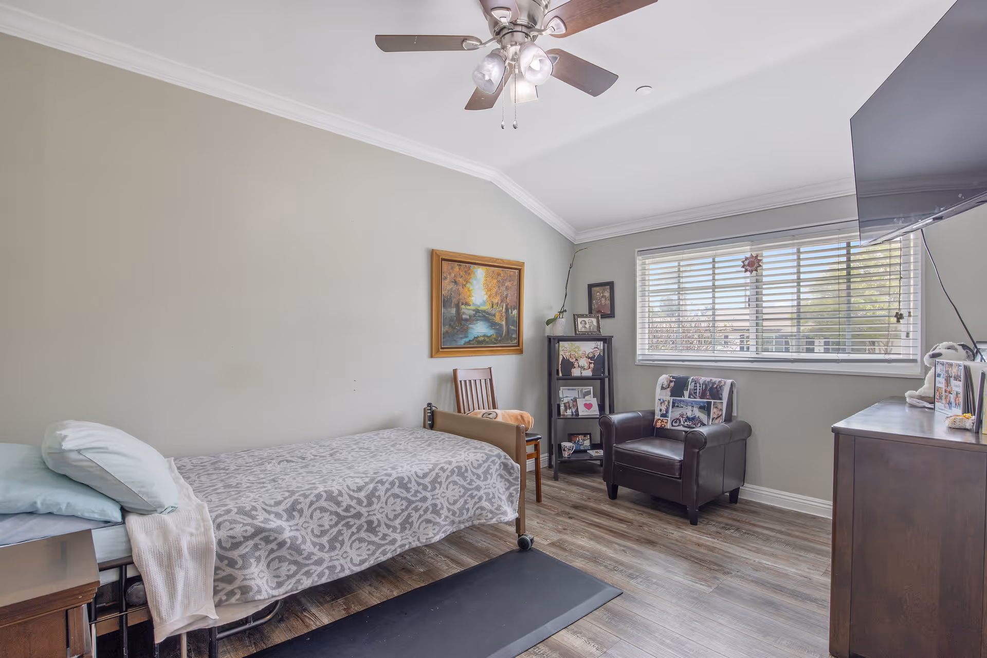 A cozy bedroom in a senior living facility with a single bed covered in a patterned gray and white blanket, a wooden chair, a small black shelving unit with framed photos, a dark leather armchair with a photo collage pillow, a large window with blinds, a ceiling fan with lights, and a wall-mounted flat-screen TV.