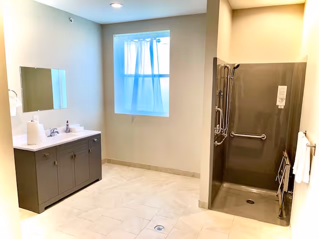 Spacious accessible bathroom with a vanity and mirror, a frosted window, tiled floor, and a roll-in shower with grab bars.