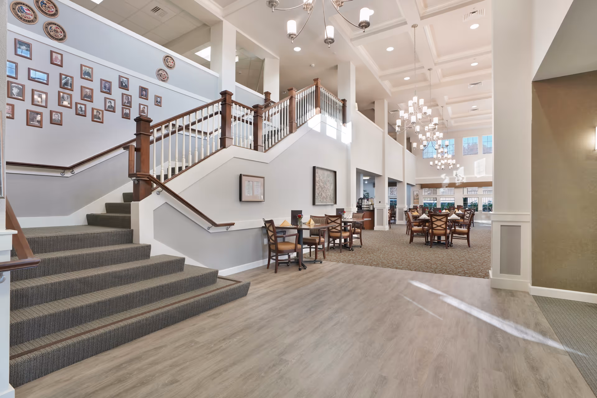Spacious interior of a senior living facility with a carpeted staircase featuring wooden handrails and white balusters. The area includes several tables and chairs arranged for dining or socializing under modern chandeliers. The walls are decorated with framed pictures and decorative plates, and large windows allow natural light to fill the space.