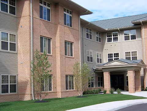 Exterior view of a senior living facility building with brick and siding facade, multiple windows, a covered entrance supported by brick columns, and a well-maintained lawn with small trees and shrubs.