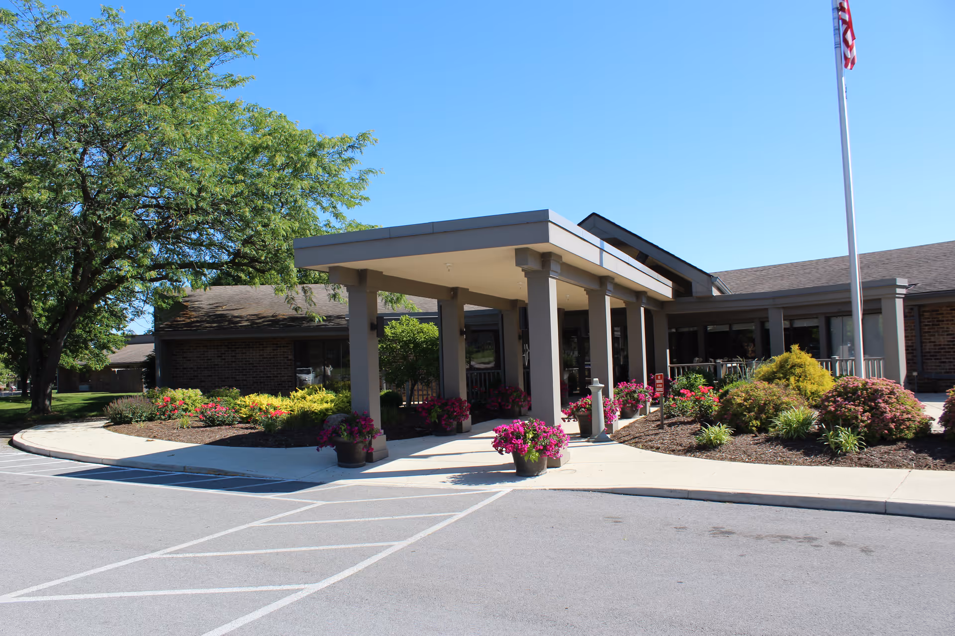 Front entrance of a senior living facility with a covered porte-cochère, columns, potted flowers and landscaped beds.