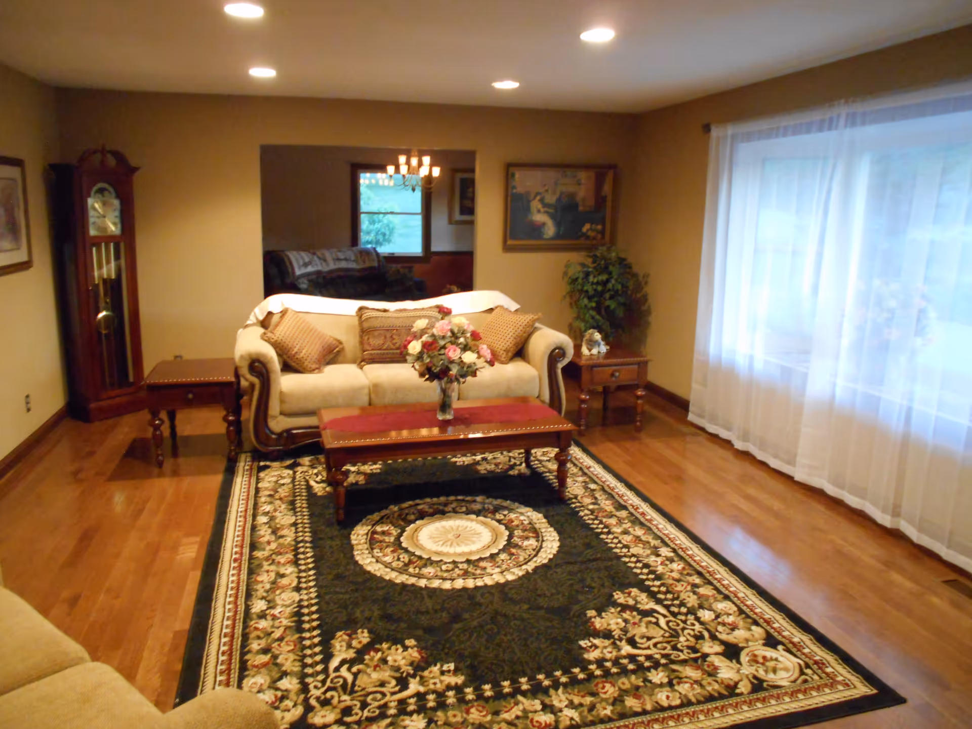 A cozy living room with a beige sofa adorned with patterned cushions, a wooden coffee table with a floral centerpiece, and matching side tables. The room features a large ornate area rug with floral designs, hardwood floors, a grandfather clock in the corner, framed artwork on the walls, and a large window with sheer white curtains allowing natural light to enter.