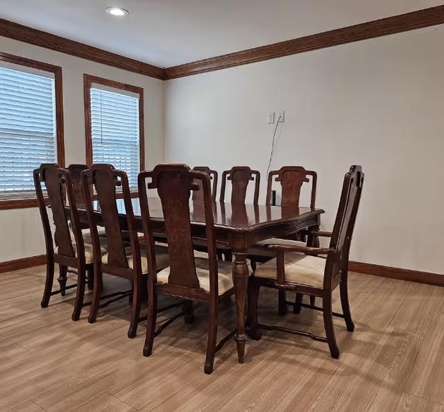 A dining room with a wooden dining table and eight matching wooden chairs with cushioned seats. The room has light-colored walls, two windows with blinds, and wooden trim along the ceiling and floor.