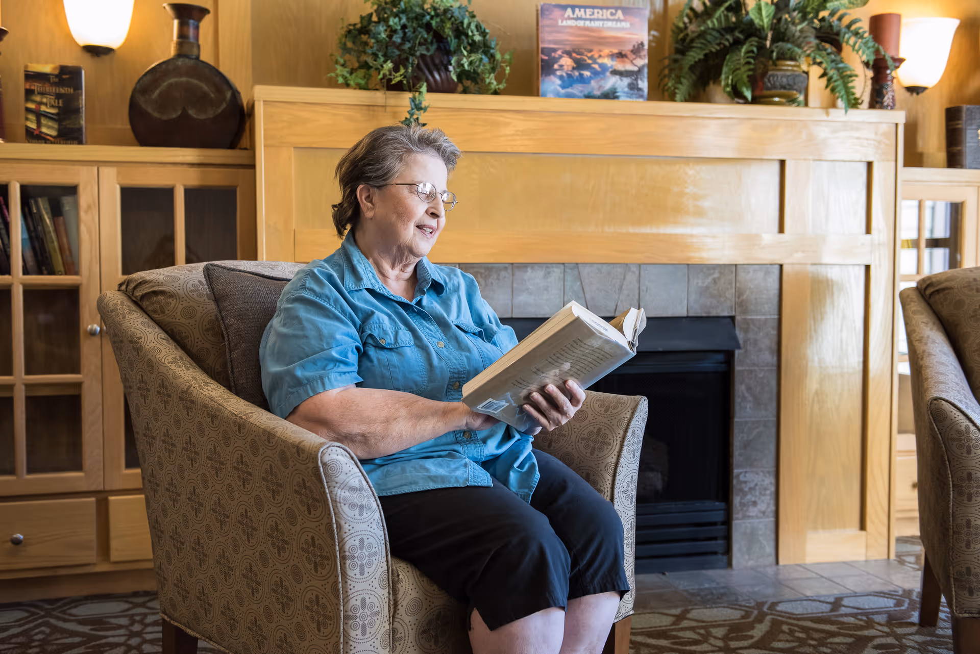 An older woman reads a book while seated in an armchair in a cozy lounge area with a fireplace and bookshelves.